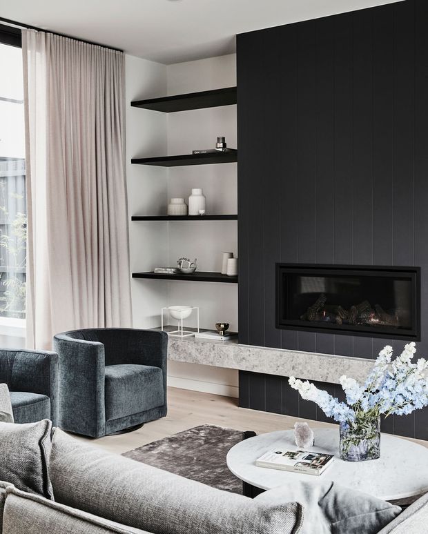 Contemporary living room with a dark vertically panelled fireplace wall, inset linear fireplace, open black display shelving, pale stone hearth, grey upholstered seating, round coffee table, and full-height curtains beside a window. The room has a refined, monochromatic palette with soft blue floral accents and natural light.