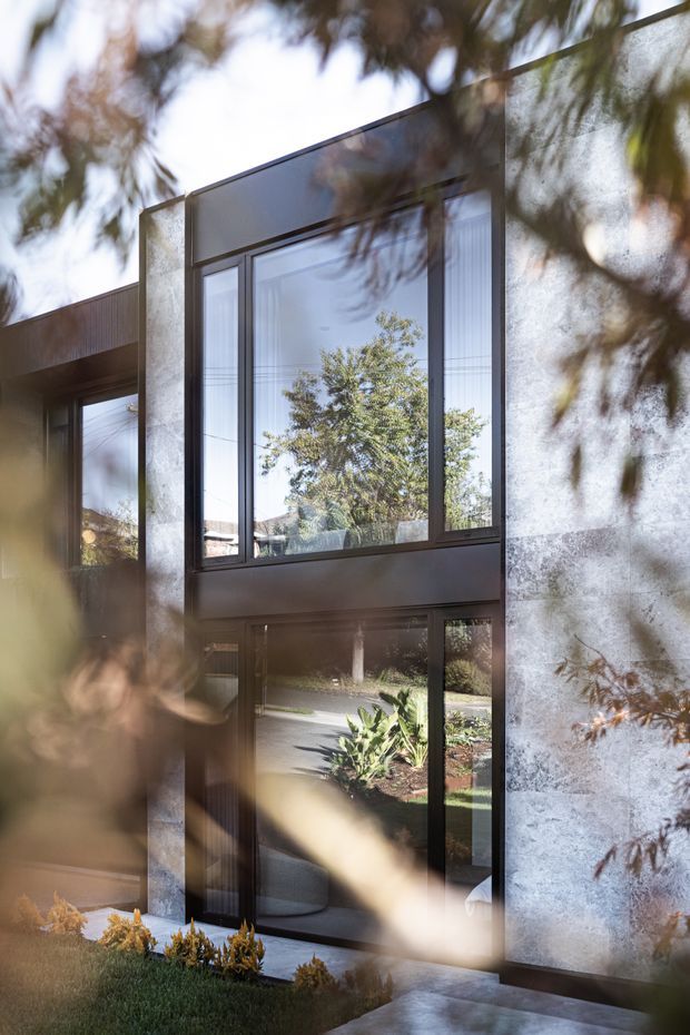 Exterior view of a contemporary multi-level residence, partially framed by out-of-focus landscaping. The facade combines expansive dark-framed glazing with a tall pale textured masonry or concrete wall, creating a strong vertical composition. Reflections in the windows show trees and the surrounding suburban landscape, while a glazed entry or lower-level opening connects to a paved path and planted garden.