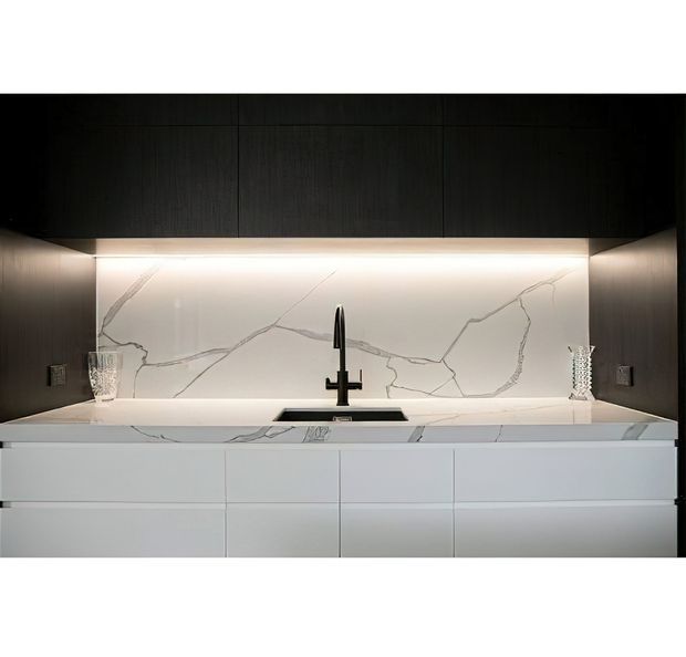 Contemporary kitchen joinery with a dark timber overhead cabinet wall, illuminated white marble-look splashback, white stone benchtop, integrated sink, and matte white lower cabinetry. The linear composition and concealed lighting create a refined, minimalist appearance.