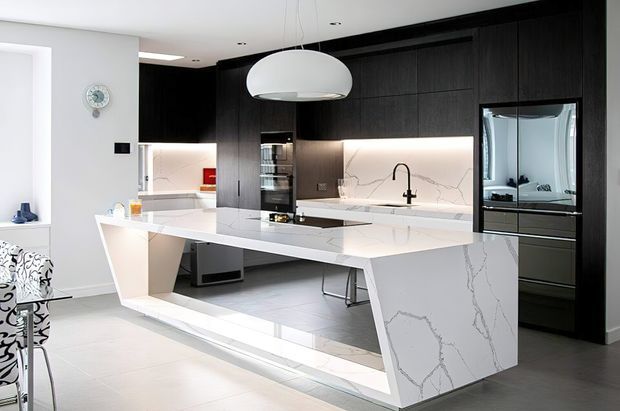 Contemporary open-plan kitchen featuring a large white waterfall-edge island with integrated seating, white veined stone surfaces, dark timber cabinetry, under-cabinet lighting, and a black-framed glass-front refrigerator. A sculptural white pendant light hangs above the island, with a dining area visible at the side.
