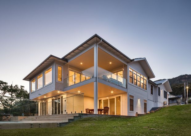 Twilight exterior view of a contemporary multi-level home on a sloped site, featuring pale horizontal weatherboard cladding, multiple gabled roof forms, extensive glazing, a covered upper balcony, and warm recessed lighting. The landscaped grass surrounds a large paved outdoor terrace and the building is set against a wooded hillside.