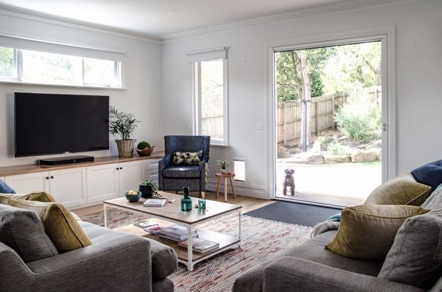 Bright, inviting contemporary living room with a large sectional seating arrangement, built-in white TV cabinetry, television, glass coffee table, patterned area rug, and generous glazing opening to a landscaped yard. The room has a neutral grey and white palette with olive and blue accents, polished timber flooring, and restrained modern detailing.