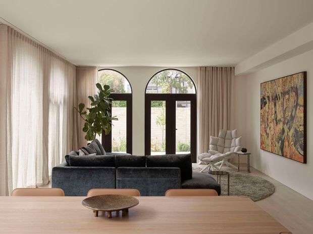 A refined open-plan living and dining area with a large timber dining table in the foreground, a dark upholstered sectional sofa, and a sculptural lounge chair. Full-height sheer and taupe curtains frame tall arched glass doors with dark frames, while a large abstract artwork and indoor tree add visual interest. The room has a warm, softly lit, contemporary feel with neutral finishes and natural textures.