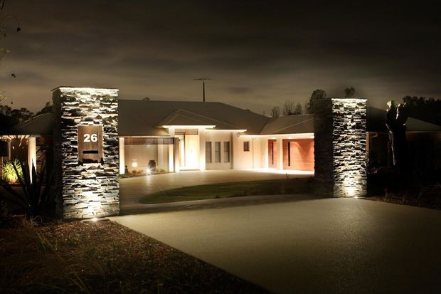 Night-time exterior view of a single-storey contemporary residence. A broad concrete driveway leads to a low-pitched roof house with strong horizontal lines, warm architectural lighting, glazed entry areas, and two illuminated stacked-stone masonry columns framing the entrance. Landscaping and dark sky surround the property.