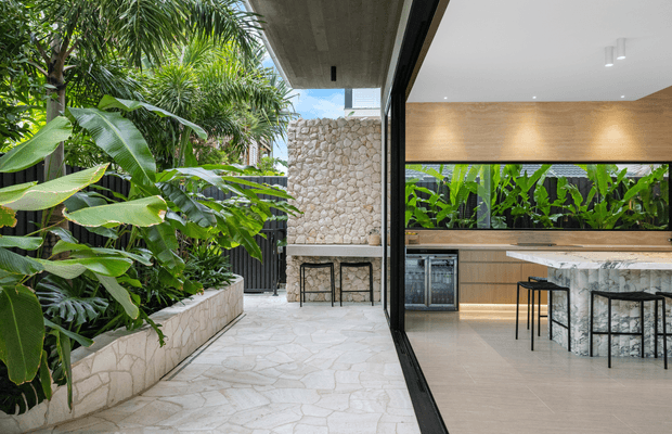 A contemporary indoor-outdoor entertaining space viewed from a leafy landscaped courtyard. The covered outdoor area has irregular pale stone paving, a raised stone planter and a stone-clad barbecue or fireplace wall. Through the large black-framed opening is a refined kitchen with timber cabinetry, a pale stone island, black bar stools, integrated refrigeration and a full-width window overlooking tropical planting. Concrete soffits and expansive glazing reinforce the connection between the kitchen and courtyard.