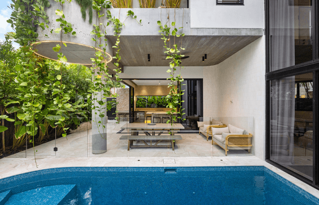 Contemporary multi-level courtyard entertaining area opening directly onto a swimming pool. The space features a covered outdoor dining zone with a timber table and bench seating, pale upholstered lounge chairs, lush climbing and potted greenery, light stone paving, a textured concrete ceiling, and large black-framed glazed doors.