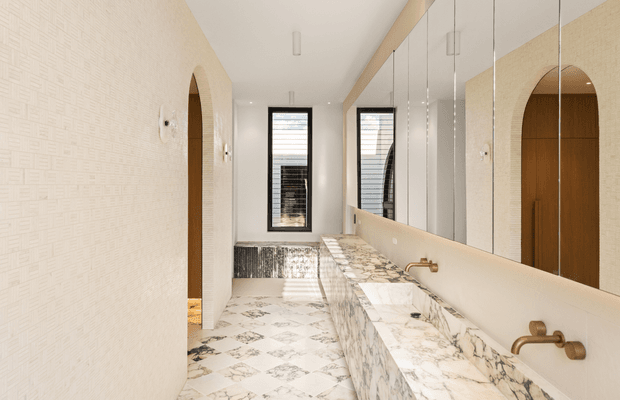 A refined, elongated bathroom with twin marble vanities, wall-mounted brass faucets, large mirrored cabinetry, patterned marble flooring, textured cream walls, and arched door openings. Slim black-framed windows bring in natural light, creating a bright, luxurious interior with strong geometric detailing.