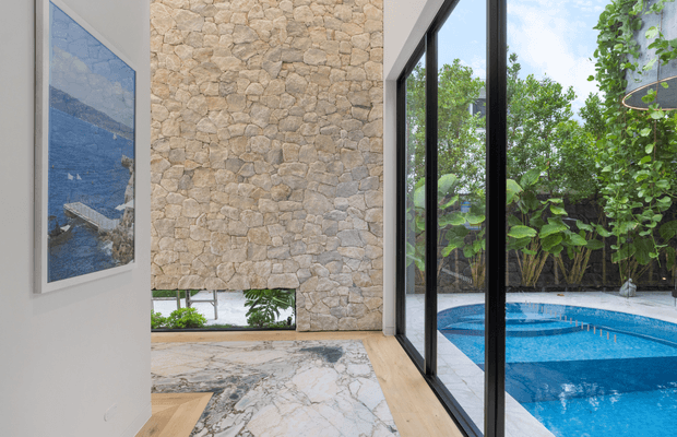 A refined contemporary interior corridor or entry space with a dramatic floor-to-ceiling natural stone feature wall, white walls, pale timber flooring, and large black-framed glass panels overlooking a bright blue swimming pool and lush garden. A large framed artwork is mounted on the left wall, while the stone paving and pool area are visible beyond the glazing.
