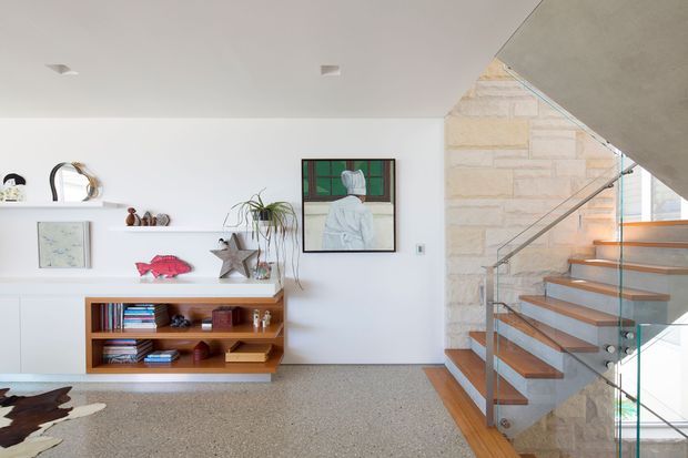 Bright contemporary interior featuring a terrazzo-finished floor, white built-in shelving and display joinery, and an open staircase with timber treads, glass balustrade, and a pale stone-clad wall. The space includes curated artwork, books, and small decorative objects, with natural light entering from the stairwell glazing.