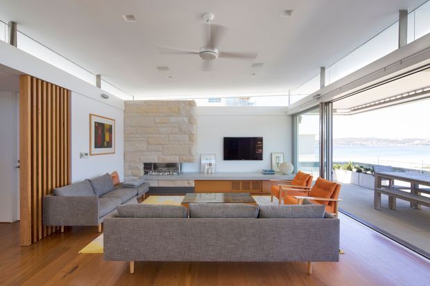 Bright, open-plan living room with floor-to-ceiling sliding glazing opening onto a waterfront deck. The room features a large grey sectional sofa, timber flooring, a stone-clad fireplace wall, timber slatted feature screening, a wall-mounted television, orange lounge chairs, and a dining table on the covered outdoor deck. The interior has a clean contemporary coastal character with abundant natural light.