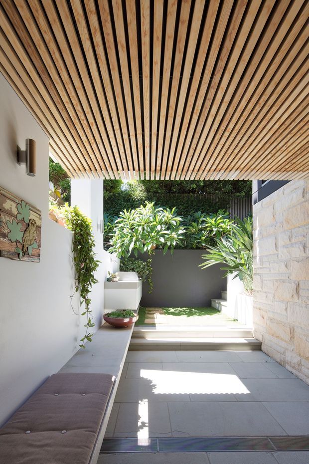 A bright, sheltered outdoor courtyard or covered terrace with large pale stone floor tiles, white rendered walls, a natural stone block feature wall, lush greenery, and a slatted timber ceiling creating a strong linear canopy. A built-in upholstered bench runs along the left wall, with steps leading to a planted garden area and a small wall-mounted basin or fixture in the background.