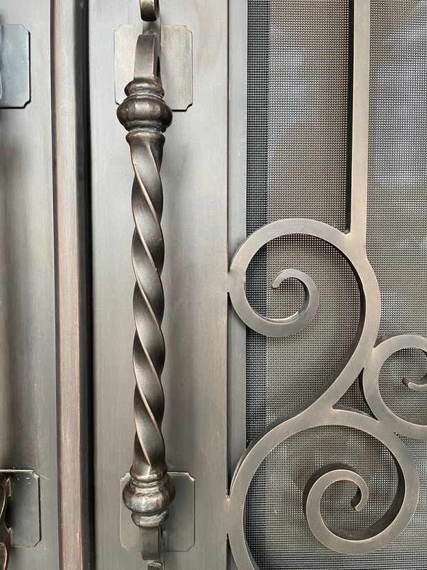 Close-up detail of an ornate metal security or decorative door grille, featuring a twisted vertical metal bar with turned finials, rectangular mounting plates, and curling scrollwork over fine mesh. The finish is dark silver-grey with a slightly aged, textured appearance.