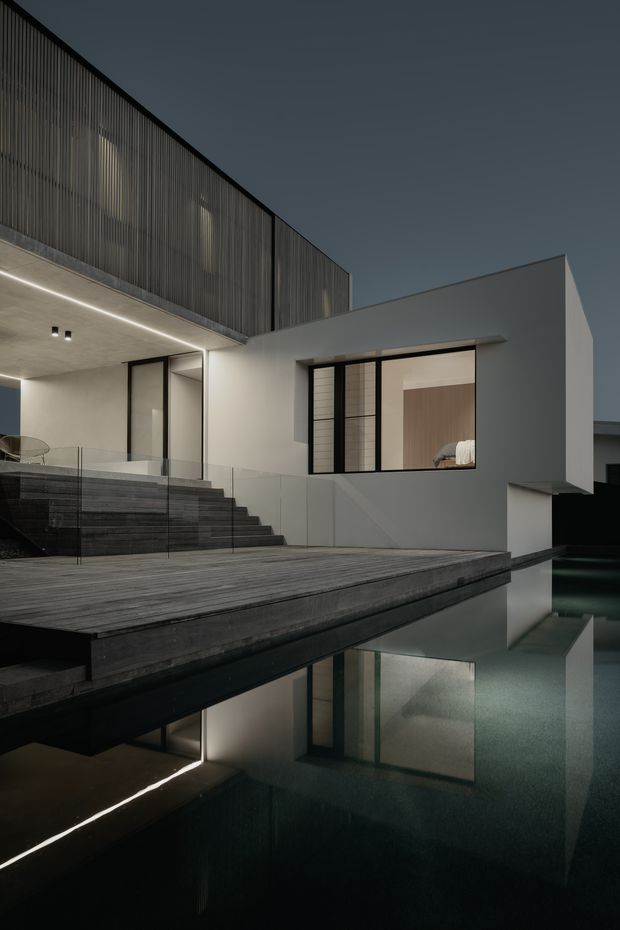 A dark, contemporary multi-level residence viewed at dusk, featuring crisp white rendered volumes, extensive black-framed glazing, a vertically screened upper level, broad timber decking, illuminated outdoor steps, and a reflective swimming pool in the foreground.