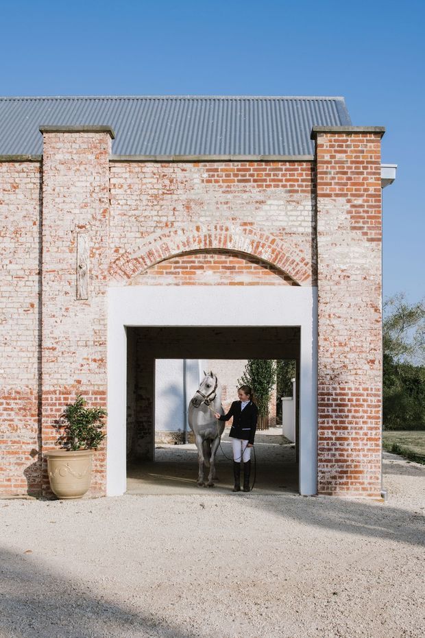 Exterior view of a historic brick stable or outbuilding with weathered whitewashed red-brick walls, a corrugated metal pitched roof, and a large arched brick detail above a rectangular gateway. A horse and rider are visible through the open passage, with gravel surfacing and a large planter in the foreground.