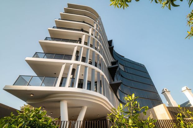 Low-angle exterior view of a contemporary multi-storey building with sweeping curved balconies, slender white structural columns, transparent glass balustrades, and a dark curved glazed facade rising behind it. Landscaping and portions of neighbouring rooflines are visible at the edges.