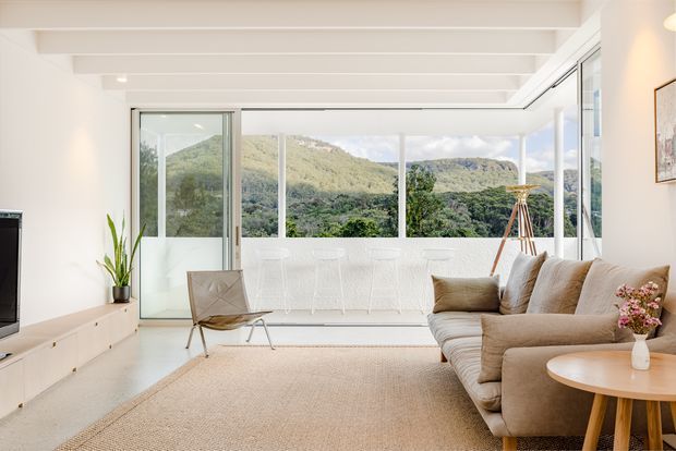 Bright, airy living room with a large beige sectional sofa, round timber coffee table, woven area rug, low white media cabinetry, sculptural lounge chair, and floor-to-ceiling glazing overlooking a green mountainous landscape. White exposed ceiling beams and slim white-framed glazing create a clean contemporary interior.