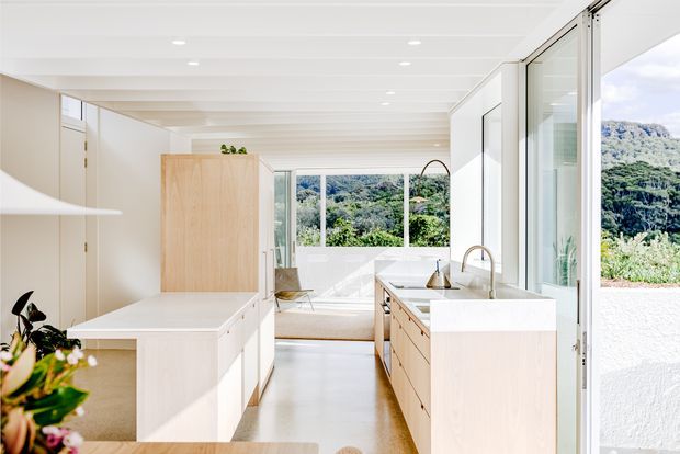 Bright contemporary kitchen with pale timber cabinetry, a large white island, white benchtops, and a stainless-steel sink and tapware. Full-height glazing and a window overlook lush greenery and a rocky hillside, while a white painted ceiling with recessed lighting creates an airy, light-filled interior.