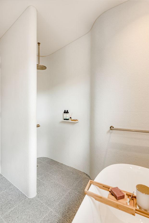 A bright, minimalist bathroom with softly curved white walls and a rounded freestanding partition. The space features a grey terrazzo-like tiled floor, a freestanding white bathtub with a wooden bath caddy, a wall-mounted brass towel rail, and a brass rainfall shower partially visible behind the partition. Fine textured white wall surfaces and warm brass fittings create a refined, spa-like atmosphere.