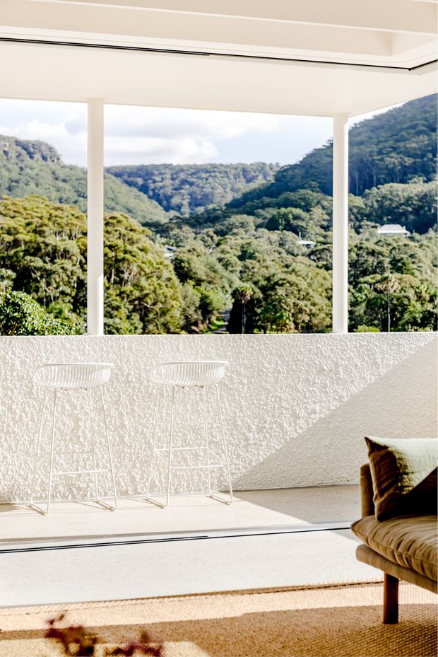 Bright, minimalist interior with expansive floor-to-ceiling glazing framing a lush, mountainous forest landscape. A textured off-white wall, slim white-framed openings, two white wire bar stools, and part of a warm brown upholstered lounge chair sit in strong sunlight above a natural woven rug.