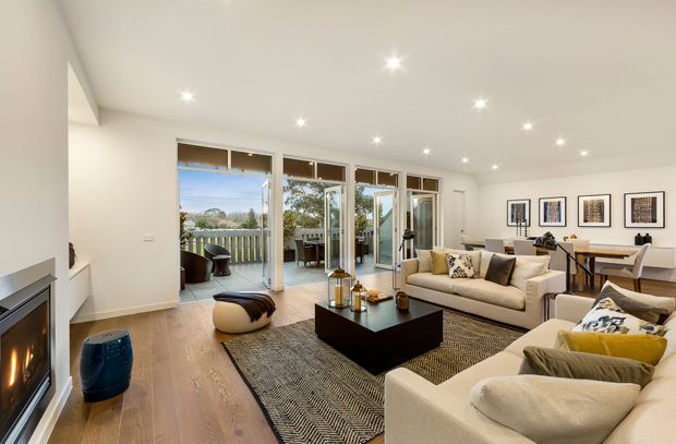 Bright contemporary open-plan living room with a fireplace, two light upholstered sofas, a black rectangular coffee table, patterned area rug, and framed artwork. Full-height glazed doors open to a balcony or terrace, bringing in abundant daylight. Timber flooring, white walls and recessed ceiling lights create a clean, inviting interior.