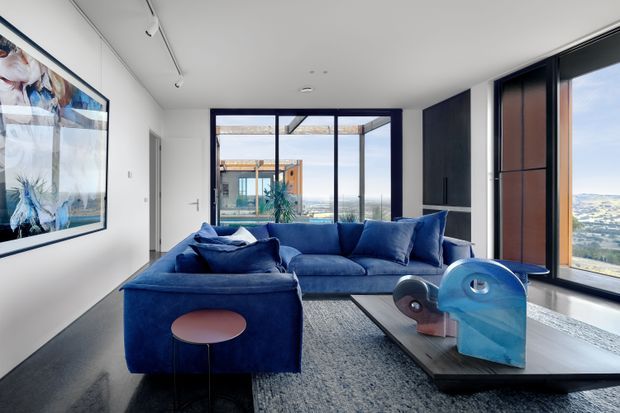 A contemporary living room with a deep navy-blue sectional sofa, sculptural coffee table, and large abstract artwork. Full-height glazing and a corner window provide expansive views of a coastal or hilly landscape, while dark-framed doors and windows contrast with white walls and a polished dark floor. Track lighting and minimal furnishings create a refined, modern interior.