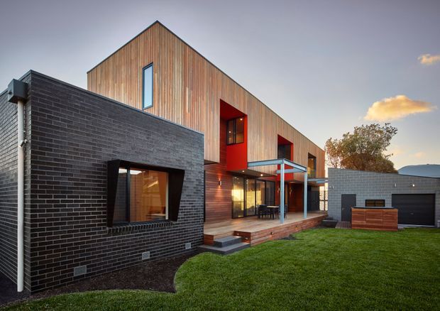 Exterior view of a contemporary multi-level residential home with a steep gabled timber-clad upper volume, dark charcoal brick lower wing, and bold red recessed entry elements. Large glazed openings connect the ground floor to a timber deck and covered outdoor area. A detached-looking garage volume, lawn, paved paths, and established planting are visible around the house.