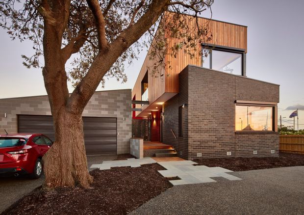 Contemporary multi-level residential exterior viewed from the driveway, featuring dark brown brickwork, warm vertical timber cladding, large windows, a projecting upper volume, and a recessed entry with red structural detailing. A flat-roofed garage wing sits to the left, while pale geometric paving leads through a sparsely landscaped front yard. A mature tree and parked car are present in the foreground.