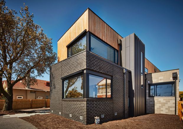 Exterior architectural photograph of a contemporary multi-level residential building. The facade combines dark brick masonry, warm vertical timber cladding, charcoal metal-framed glazing, and a lighter brick side wing. A tall gabled timber-clad volume rises above a lower brick section, with a prominent vertical metal-clad feature between the main building and side wing. Established suburban housing and a mature tree are visible around the property.