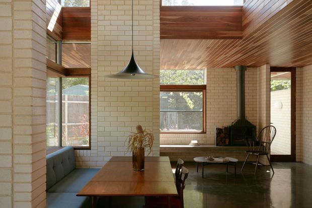 Sunlit open-plan dining and living space with cream brick walls, polished concrete flooring, warm timber-lined ceiling sections, high clerestory glazing, and dark timber-framed windows. A timber dining table with built-in upholstered bench seating occupies the foreground, while a compact lounge area, sculptural pendant light, fireplace, and occasional chairs sit beyond. The architecture has strong mid-century-inspired detailing with exposed brick, timber, and abundant natural light.