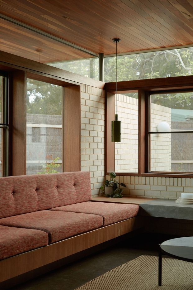 A warm mid-century-inspired living room with a built-in upholstered sectional beneath a richly timber-lined ceiling. Dark timber window and door frames surround extensive glazing, while cream brick walls, a concrete or dark stone-like benchtop, and a suspended cylindrical pendant define the space. A woven rug, low round coffee table, and indoor plant complete the restrained natural interior.