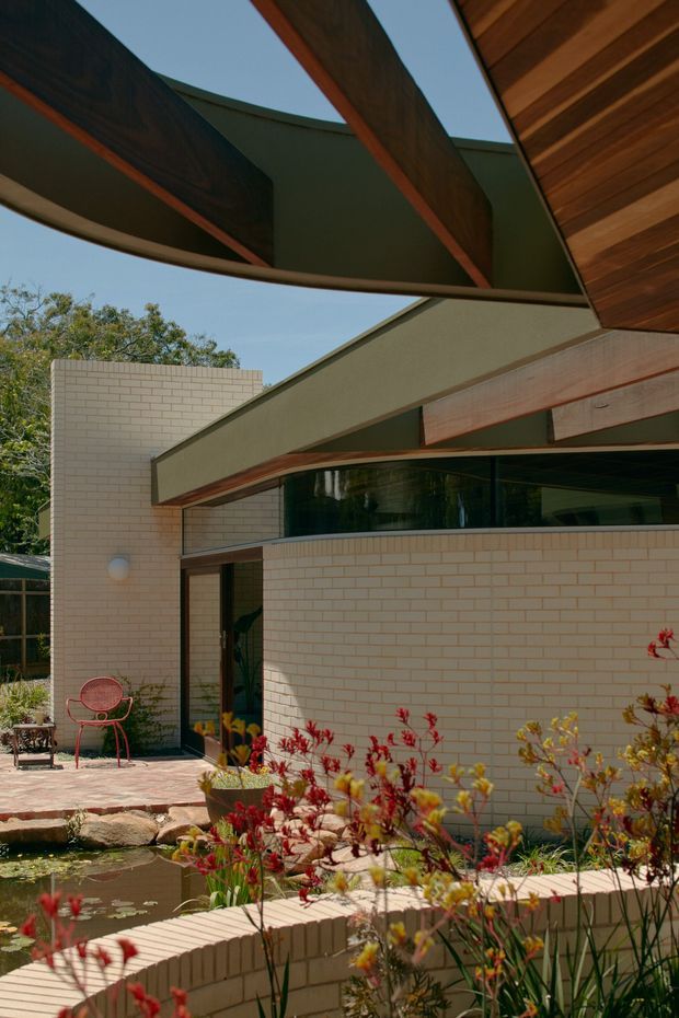 Exterior view of a low-rise contemporary residence with pale brick walls, expansive dark glazing, projecting timber-framed roof forms, and pronounced exposed structural beams. A landscaped courtyard with a shallow pond, paving, and flowering plants occupies the foreground.