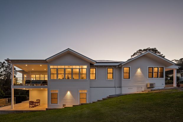 Exterior dusk view of a multi-wing, multi-storey home on a sloped grassy site. The house has light horizontal weatherboard cladding, multiple pitched gabled roofs, extensive warm-lit windows, and a large covered outdoor terrace or balcony at the left.