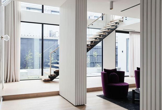 Bright contemporary double-height interior with floor-to-ceiling glazing, a floating open-riser staircase, white vertically fluted wall panels or columns, pale timber flooring, and a deep purple lounge chair on a dark rug. The space appears to be an open-plan living area with a mezzanine or upper-level void, overlooking a small courtyard.
