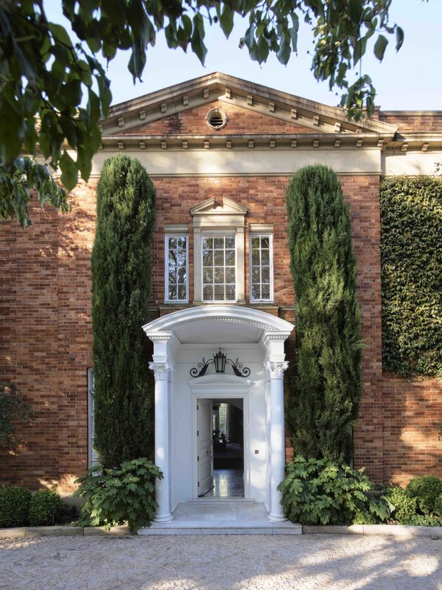Formal front facade of a stately red-brick residence, framed by mature landscaping. The symmetrical elevation features a classical pediment with dentil detailing, stone trim, a circular oculus, tall sash windows, and an elaborate white portico entrance with columns, curved canopy, decorative capitals, and wrought-iron detailing. A paved approach and clipped evergreen planting reinforce the grand, traditional character.