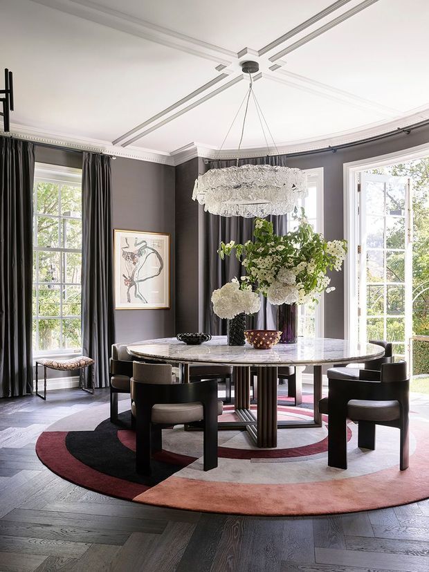 A refined formal dining room with charcoal-grey walls, elaborate white decorative ceiling mouldings, tall curtained windows and French doors, and a large sculptural crystal chandelier. A round marble dining table with dark sculptural legs is surrounded by upholstered dining chairs and sits on a circular abstract rug over dark herringbone timber flooring. Floral arrangements and framed artwork add decorative accents.