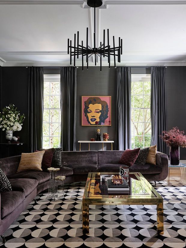 A dramatic, richly layered living room with charcoal walls, white ornate ceiling mouldings, tall curtained windows, and a sculptural black multi-arm chandelier. A deep curved sectional sofa surrounds a glass-topped brass coffee table, set over a bold black, white, and grey geometric rug. Decorative cushions, floral arrangements, a console table, and a framed pop-art portrait create a glamorous, eclectic atmosphere.
