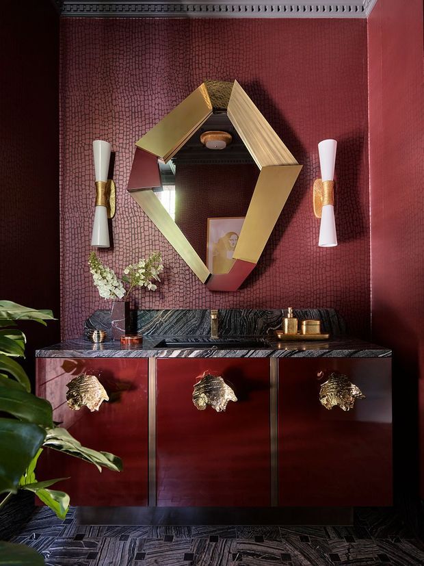 A dramatic, richly decorated interior vignette featuring a dark red console cabinet with glossy red fronts and ornate gold animal-head pulls. The cabinet sits against a deep burgundy textured wall, topped with a dark veined stone surface and brass accessories. Above it hangs an angular geometric mirror with gold-toned framing, flanked by sculptural wall sconces. A leafy plant enters from the foreground, and the dark patterned floor and ornate ceiling trim reinforce the opulent, theatrical character.