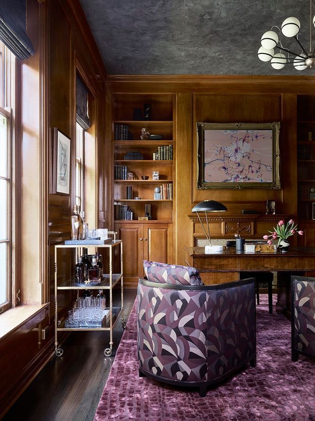 A richly detailed, traditional wood-panelled study or library with floor-to-ceiling built-in bookshelves, a carved wood desk, upholstered lounge chairs, a drinks trolley, framed artwork, tall windows with blinds, and a dramatic multi-globe chandelier. The room has a dark, warm and highly layered character, with patterned purple upholstery and carpet contrasting against the timber joinery.