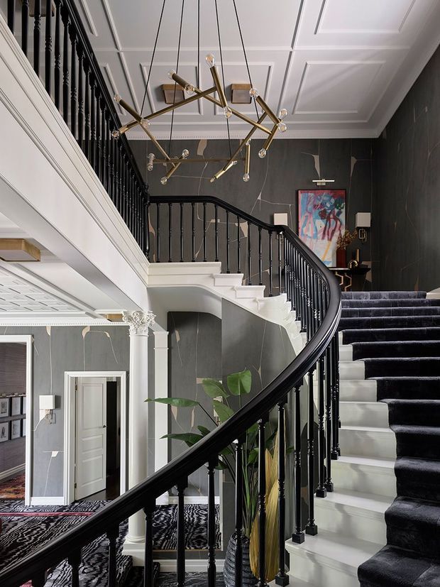 A dramatic, double-height residential stair hall with a sweeping curved staircase, glossy black balustrade, white stair treads and ornate white classical trim. Dark charcoal wall panelling, a coffered white ceiling, patterned carpet runners, a sculptural brass-and-glass chandelier, framed artwork and indoor plants create a refined, theatrical interior.