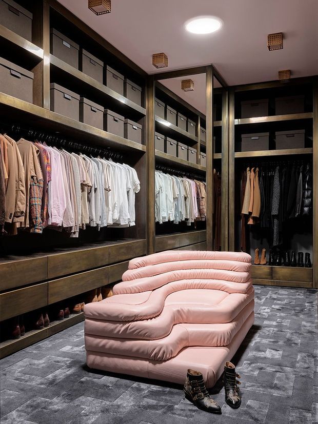 A luxurious walk-in wardrobe with dark timber or dark-stained built-in shelving, drawers, hanging rails, and neatly arranged clothing and shoes. A large sculptural blush-pink upholstered lounge sits centrally on a grey patterned carpet, while mirrors and warm decorative ceiling lights enhance the room.