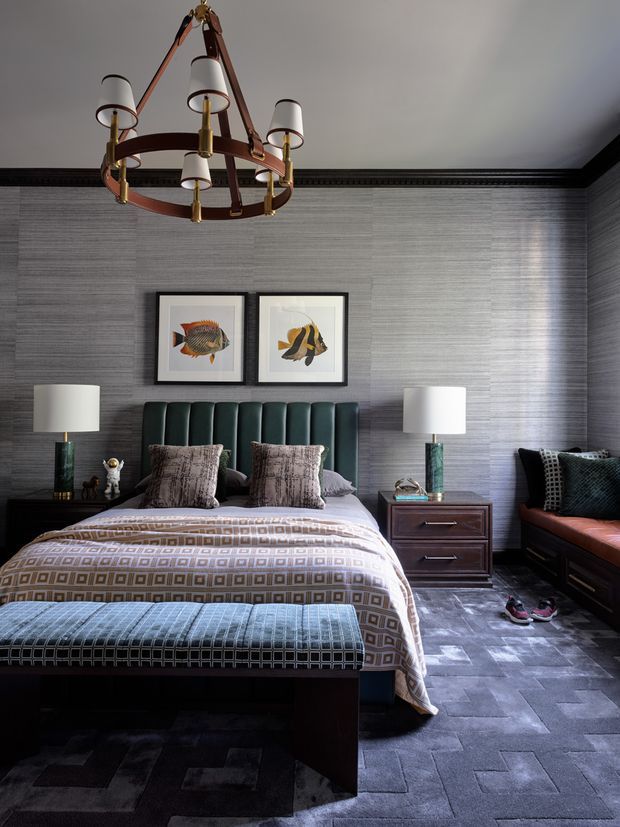 A richly layered bedroom with a dark, tailored aesthetic. The room features a tall channel-tufted deep green upholstered bedhead, patterned geometric bedding and a matching upholstered bench, dark timber bedside furniture, matching table lamps, framed fish artwork, and a substantial leather-and-brass chandelier. Charcoal textured wallcovering, dark patterned carpet, black trim, and a built-in window seat or bench create a moody, refined atmosphere.