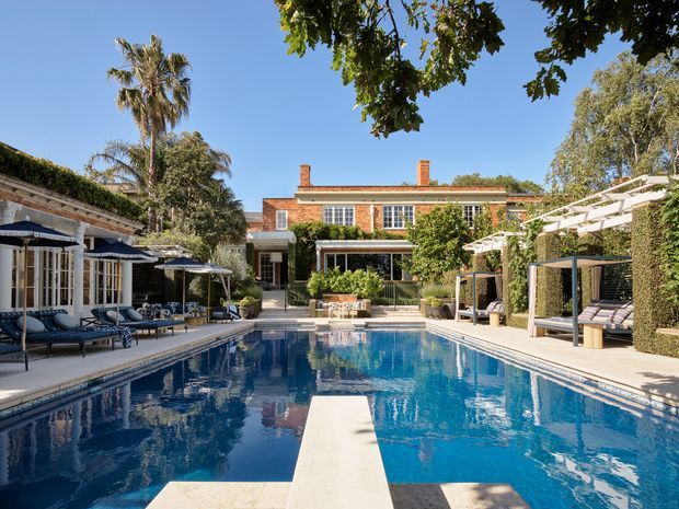 A spacious landscaped courtyard centered on a large rectangular swimming pool with a diving board. The pool is surrounded by pale stone paving, shaded lounge furniture, umbrellas, pergola structures, and clipped hedges. A substantial red-brick multi-storey house with white-trimmed windows and covered outdoor seating forms the backdrop, with mature palm and leafy trees around the property. Bright, upscale residential outdoor setting.