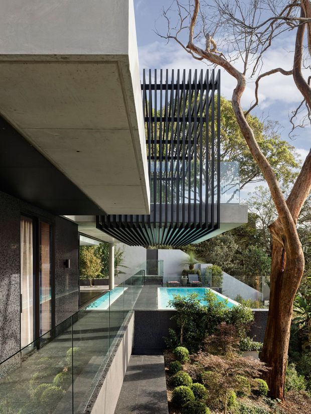 A contemporary multi-level residential exterior viewed from a terrace, featuring strong cantilevered concrete slabs, dark textured walls, extensive glass balustrades, and a dramatic black vertical louvre screen around an upper-level balcony. A turquoise swimming pool occupies the courtyard terrace, surrounded by landscaped planting and mature trees.