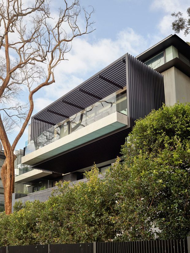 Exterior view of a contemporary multi-storey residence, photographed from below. The building features strong horizontal cantilevered balcony volumes, dark vertical metal screening, glass balustrades, a projecting pergola with closely spaced slats, and light textured wall panels. Dense landscaped hedging and mature trees partially obscure the lower level.