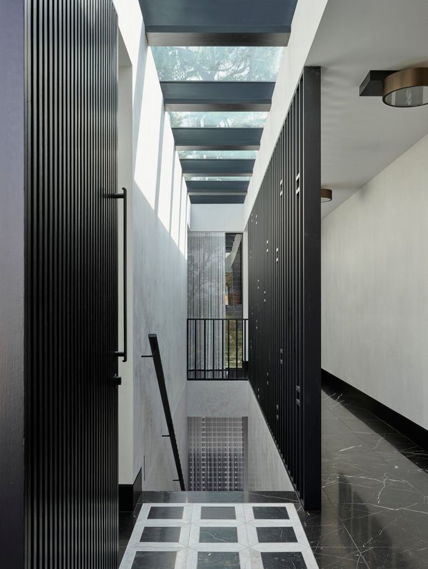 A narrow, sophisticated interior stairway or landing corridor with black vertically ribbed architectural screens and doors, white plaster walls, dark stone flooring, and a dramatic glazed overhead skylight framed by exposed dark beams. A patterned geometric stone floor marks the foreground, while a black metal balustrade overlooks a lower level.