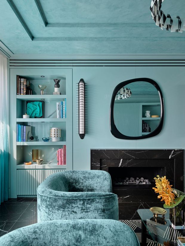 A richly styled living room in a deep teal and black palette. The room features a sculptural teal velvet armchair and ottoman, a black marble fireplace surround, built-in teal shelving with books and decorative objects, fluted wall cabinetry, a rounded black-framed mirror, and a dramatic black-and-white pendant light reflected in the mirror. The ceiling has recessed geometric detailing, while dark stone flooring and glossy finishes create a sophisticated, moody atmosphere.