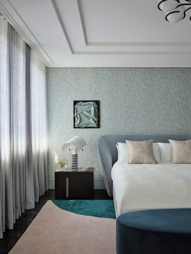 A refined bedroom interior with a curved upholstered bed in blue-grey, white bedding, and neutral decorative cushions. The room features a textured pale blue wall, full-height sheer curtains, dark flooring, a black bedside cabinet, sculptural table lamp, layered curved rugs, and a recessed tray ceiling with a contemporary pendant light. The palette is cool and elegant with soft contrasting textures.