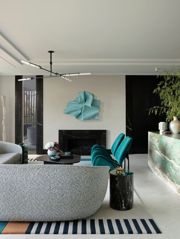 A sophisticated contemporary living room with a large curved grey sofa in the foreground, vivid teal sculptural lounge chairs, a black marble fireplace surround, and a pale stone or marble bar/console at right. The room features light flooring, a dark ribbed feature wall, abstract turquoise wall art, decorative vases and greenery, and a striking black linear pendant light.