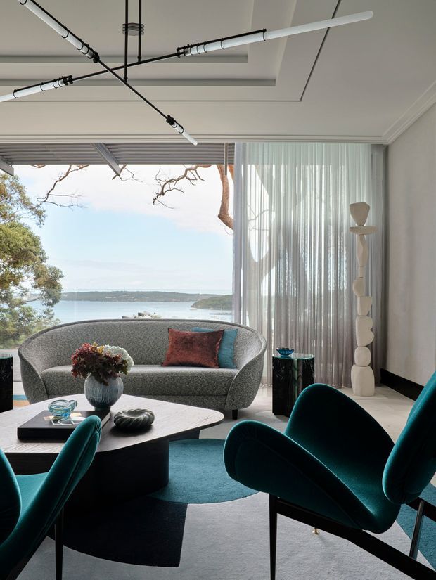 Refined living room with a sculptural grey sofa and deep teal lounge chairs arranged around a low dark coffee table. The room has a large glazed opening with sheer curtains overlooking a waterfront landscape, a patterned teal rug, decorative objects, and a contemporary ceiling with exposed linear lighting and recessed detailing.