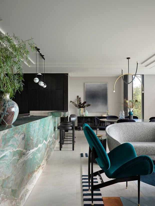 A sophisticated open-plan interior combining a dramatic veined green stone island and bar with a dining and living area. The space features black fluted wall panelling, pale flooring and walls, sculptural pendant lighting, teal velvet dining chairs, a textured curved sofa, and contemporary artwork and styling. Large glazing brings in natural light and greenery is used as an interior accent.